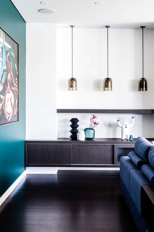 A bright contemporary living room featuring a dark timber built-in sideboard and shelving, a deep teal feature wall with framed artwork, three suspended pendant lights, dark timber flooring, and a blue upholstered sofa. Decorative objects sit on the sideboard, creating a refined, high-contrast interior.