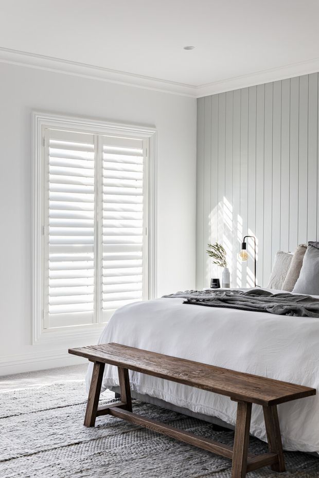 Bright, softly styled bedroom with a white upholstered or dressed bed, layered grey bedding and pillows, a rustic timber bench at the foot of the bed, white plantation shutters, and a vertical white wall-panel feature behind the bed. Natural daylight creates a calm, airy atmosphere, with a textured grey rug and minimal bedside styling.