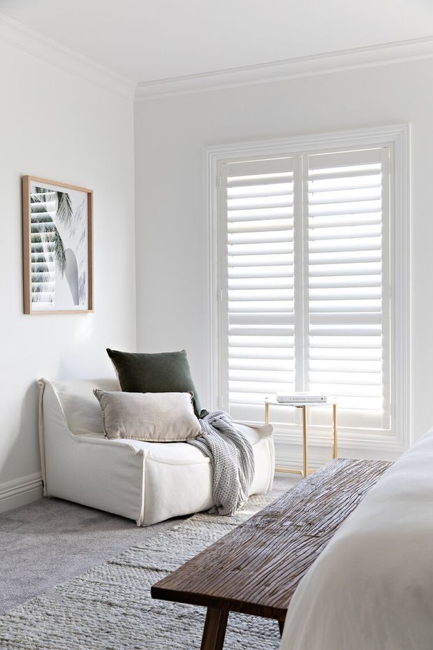 Bright, refined bedroom corner featuring a white upholstered armchair with layered cushions and a draped knitted throw beside a large shuttered window. A slim gold-toned side table, framed monochrome artwork, soft grey carpet, textured area rug, and a dark timber bench at the foot of the bed complete the calm neutral interior.