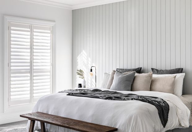 Bright, softly styled bedroom with a large upholstered bed dressed in white bedding and layered neutral pillows. The room features white plantation shutters, a vertical white panelled feature wall, pale walls with decorative cornice, and a rustic dark timber bench at the foot of the bed. Natural light creates a calm, airy atmosphere.