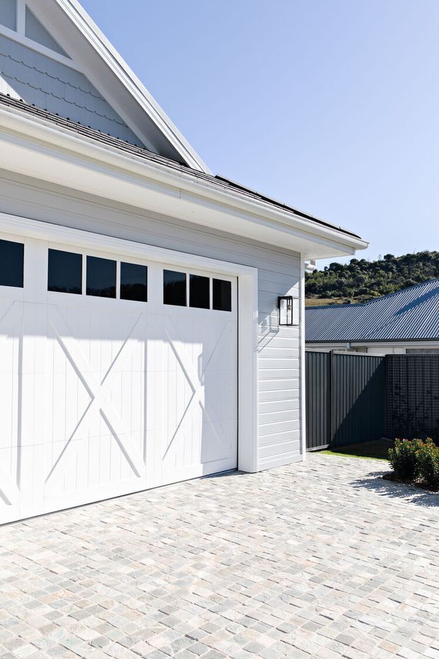 Bright exterior view of a white weatherboard garage with a steep gabled roof, decorative barn-style garage door, small upper window panels, and white trim. The foreground is a broad paved driveway of light grey and beige stone pavers, with a dark metal fence and another corrugated-roof structure in the background.