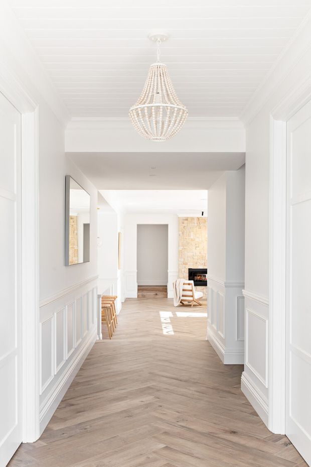 Bright, elegant residential hallway with white painted walls, detailed panelled wainscoting, a white beadboard-style ceiling, and warm pale timber herringbone flooring. A decorative beaded pendant chandelier hangs centrally, with a large wall mirror and glimpses of a relaxed living area and fireplace beyond.