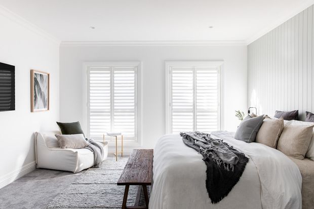Bright, refined bedroom with a large upholstered bed, layered neutral bedding, a timber bench, a lounge chair, plantation shutters, and a white vertical-panel feature wall. The room has abundant natural light and a soft grey, white, and taupe palette.