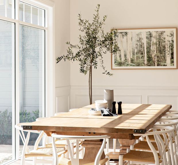 Bright, refined dining room with a long natural timber dining table surrounded by white and woven chairs. Floor-to-ceiling glazed doors or windows bring in abundant daylight, while white wall panelling, a framed forest artwork, and an indoor olive tree create a calm, natural atmosphere.