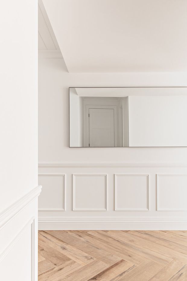 Bright, refined interior hallway or landing with white painted walls, traditional decorative wall panelling and mouldings, a large horizontal mirror, and light natural timber herringbone flooring. The mirror reflects a white paneled door and adds depth to the narrow space.