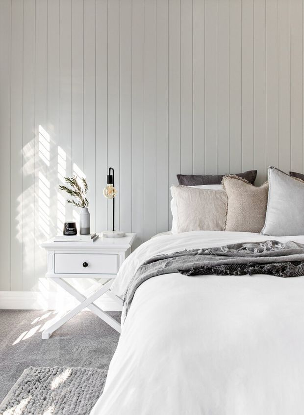 Bright, calm bedroom scene with a white upholstered bed dressed in layered white and grey bedding, positioned beside a small white bedside table with crossed-leg supports. The room features pale vertical wall panelling, light grey carpet, neutral pillows and throws, a slim black floor lamp, and simple minimal decor. Strong sunlight creates angled highlights and shadows across the wall and floor.