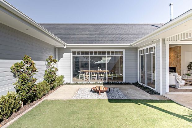 A bright landscaped courtyard enclosed by a light-grey weatherboard home. The courtyard features a small paved patio with a central fire bowl, white gravel edging, clipped planting, and a neat lawn. Large white-framed glazed openings connect to an interior dining or living space, while the roof is clad in grey shingles and the covered veranda includes decorative white detailing.