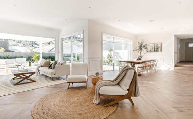 Bright, spacious open-plan living and dining area with large windows, pale timber flooring, white decorative wall panelling, and light neutral furnishings. A seating area with armchairs and sofas opens onto a dining zone, with an entry hallway visible at right.
