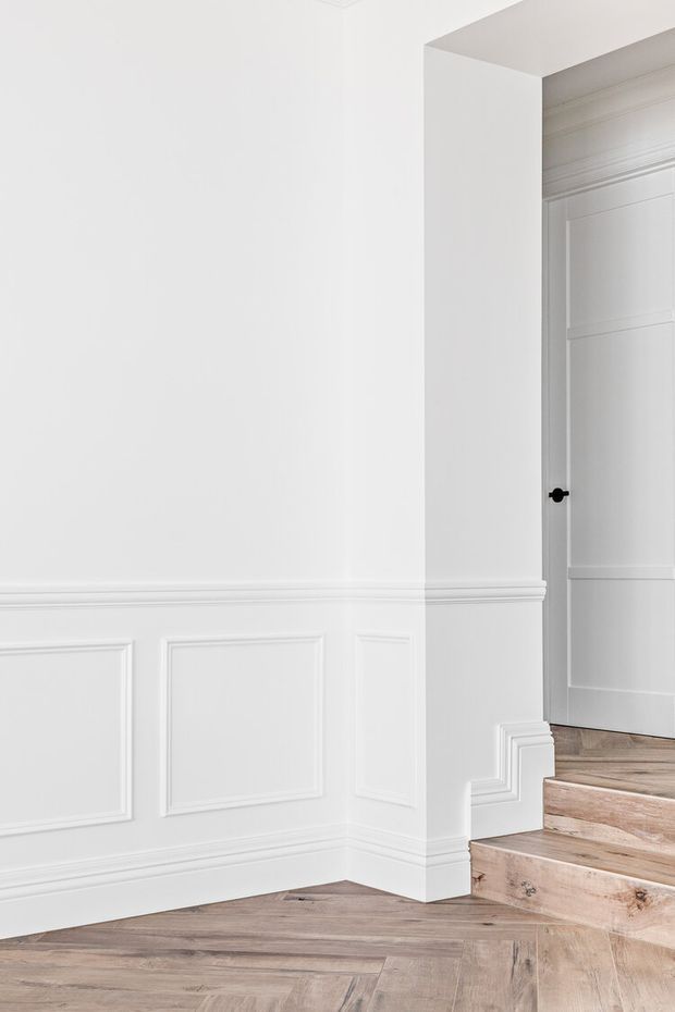 A bright, refined interior hallway or stair landing featuring white painted walls with extensive decorative wainscoting, moulded dado rails and skirting boards. A white paneled door with a black knob opens at the top of a short set of timber stairs. The floor is finished in warm, natural-toned herringbone timber flooring, continuing onto the stair treads. The composition is minimal and monochromatic with traditional detailing.
