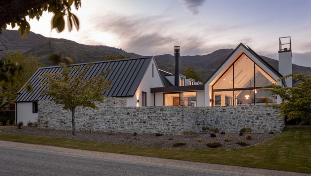 Exterior view of a contemporary mountain home at dusk, composed of steep gabled volumes with dark standing-seam metal roofing, pale stone walls, extensive glazed gable frontage, and warm interior lighting. A low dry-stone boundary wall and landscaped front garden sit between the house and the street, with rugged mountains forming the background.