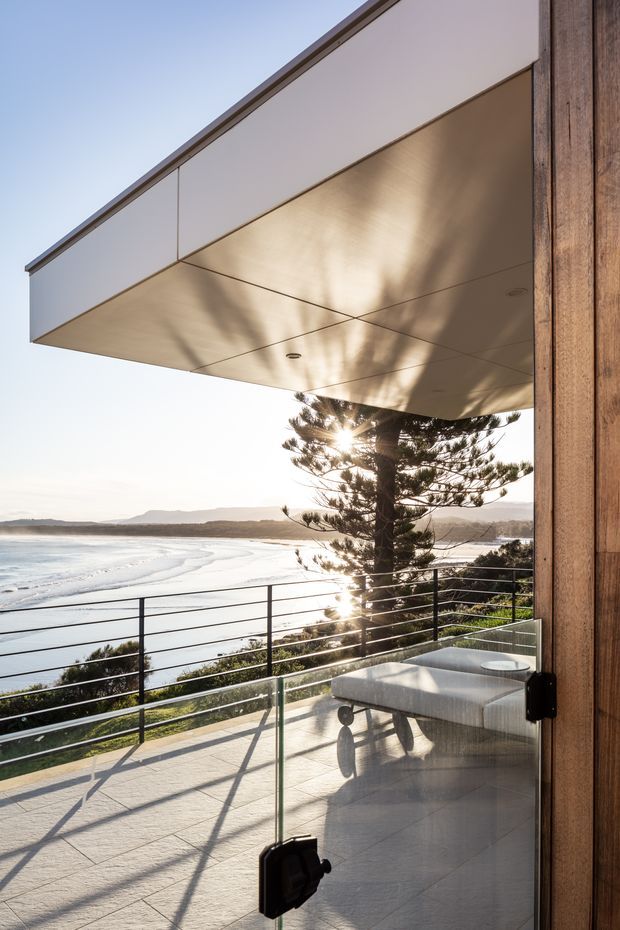 A contemporary coastal balcony or terrace viewed from beside a timber-framed glass door. The broad cantilevered roof canopy has a smooth pale underside with recessed downlights, while a glass balustrade and dark horizontal metal railing frame an expansive ocean and beach outlook. A built-in pale upholstered bench sits at the edge, with a Norfolk Island pine and coastal landscape beyond.