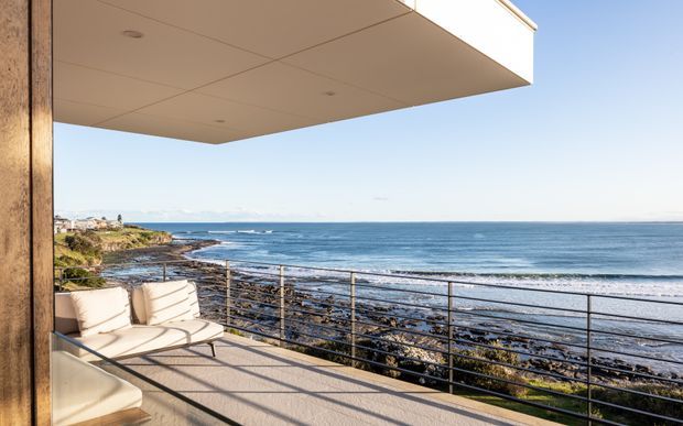 A bright coastal terrace or balcony extending over a rocky shoreline, with a minimalist metal balustrade, pale outdoor lounge seating, timber-clad structural post, and a broad projecting soffit. The ocean and distant coastline dominate the view, creating an open waterfront setting.