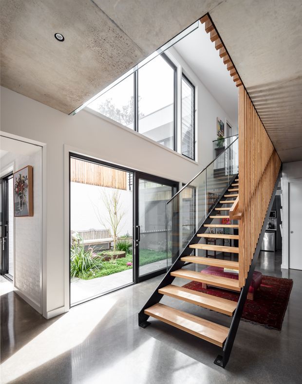 Bright contemporary interior featuring an open-riser timber and black steel staircase with clear glass balustrades, rising through a double-height void. The space has polished concrete floors and ceilings, large aluminium-framed glazing, a timber slat screen, and a small landscaped courtyard visible through the doors.