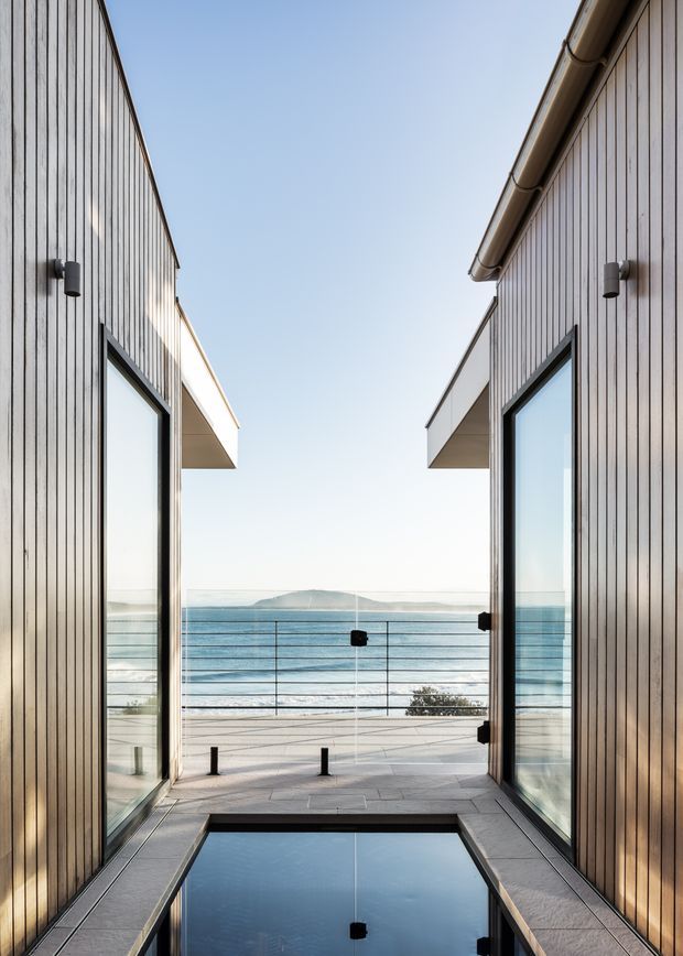 A narrow, sheltered outdoor courtyard or terrace between two vertically clad building volumes, opening toward the ocean. Full-height glazed doors, dark-framed glazing, a shallow reflecting pool, stone paving, glass balustrading, and simple wall-mounted lights create a clean contemporary coastal composition.