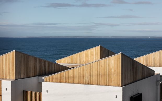 Exterior architectural view of a cluster of angular, gabled roof forms clad in vertical warm timber boards above white rendered walls. The geometric composition is set directly against a broad ocean horizon under a lightly clouded sky.
