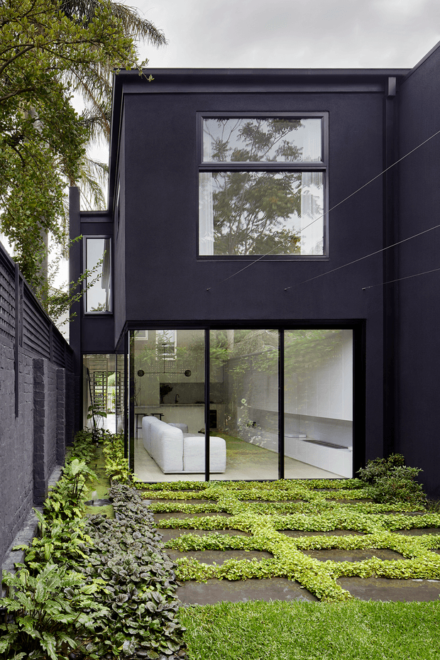 A narrow landscaped courtyard beside a tall, dark-clad contemporary house. The two-storey facade features large black-framed windows and floor-to-ceiling glazing opening onto a stepped garden of concrete pavers densely planted with low groundcover and leafy borders. A minimalist white sofa and interior shelving are visible through the glazing, while a dark masonry boundary wall and mature vegetation frame the space.
