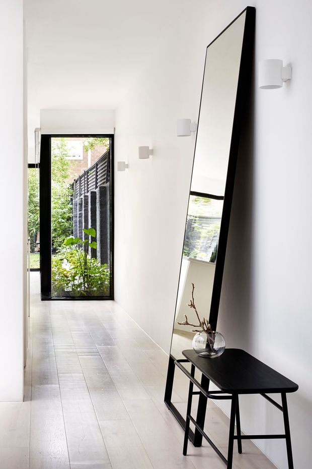 A bright, narrow minimalist hallway with white walls, pale timber-look tiled flooring, black-framed glazed door at the end, and a large angular black-framed leaning mirror above a slim black console table. Simple white wall sconces line the corridor, while greenery is visible through the glazed door and reflected in the mirror.