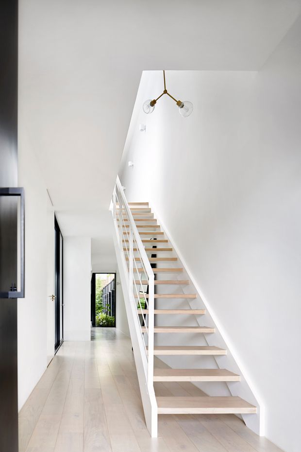 A bright, minimalist interior hallway dominated by an open-riser staircase. The staircase has pale timber treads, white stringers and balustrades, and rises alongside a tall white wall toward an upper landing. Pale timber flooring, white plaster walls, black-framed glazing, and a simple brass branching pendant light create a clean, airy Scandinavian-contemporary atmosphere.