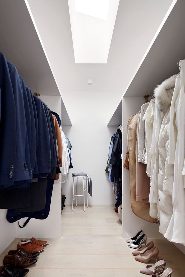 A bright, narrow walk-in wardrobe with clothing hanging on both sides, shoes arranged along the floor, pale timber or wood-look flooring, white built-in shelving, and a large rectangular skylight or ceiling light well. The space has a clean, organised, contemporary appearance.