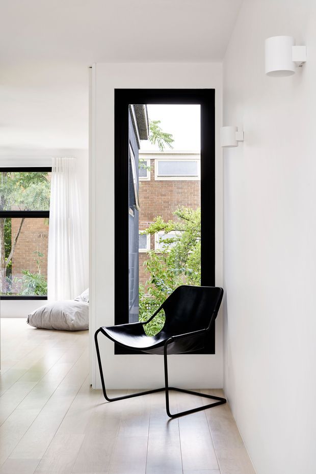 Bright minimalist interior corner with a black leather sling chair positioned beside a tall black-framed window. Pale timber flooring and white walls create a clean, airy hallway or landing, while an adjacent bedroom with a low bed and sheer curtains is visible to the left. Simple white wall sconces provide restrained architectural lighting.