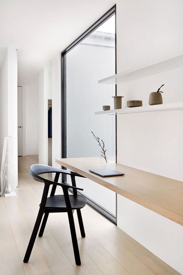A bright, minimalist home workspace arranged along a large black-framed internal or external glazed wall. A pale timber desk with integrated white cabinetry runs beneath floating white shelves displaying understated ceramic objects and a branch. A black sculptural armchair and laptop sit at the desk, while pale timber flooring, white walls, and a glimpse of a hallway and wardrobe create a clean, airy composition.