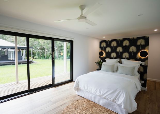 Bright contemporary bedroom with a large upholstered bed dressed in white linens, a dark feature wall with repeating gold botanical motifs, and twin circular wall lights. Full-height black-framed sliding glass doors open the room toward a timber deck and leafy lawn. The room has pale timber flooring, a textured woven rug, recessed ceiling lights, and a white ceiling fan.