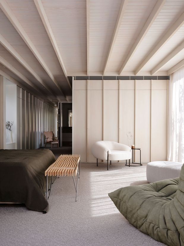A serene, contemporary bedroom with a partially visible bed, sculptural lounge chairs, a low slatted bench, soft carpet, full-height curtains, and pale timber wall panelling. The room is defined by a dramatic exposed-beam ceiling with light timber boards and beams, while sunlight filters through the sheer curtains. A dark timber-toned passage or adjoining area is visible in the background.