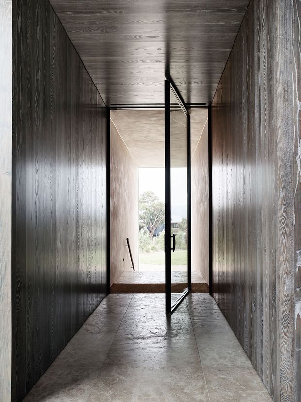 A narrow, dramatic entrance corridor lined with dark vertically grained timber wall and ceiling panels. Large-format grey stone or concrete-look flooring leads to a full-height black-framed glazed door opening toward a bright landscaped exterior. The restrained palette, strong linear perspective, and concealed detailing create a minimalist, refined architectural interior.