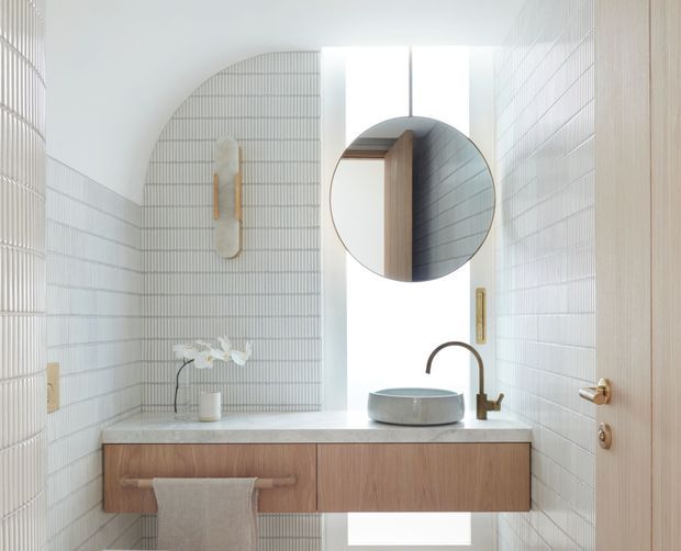 Bright, minimalist bathroom vanity with a floating timber cabinet, pale stone benchtop, round vessel basin, brushed bronze tapware, white vertically lined wall tiles, arched tiled feature wall, round mirror, and concealed natural light.