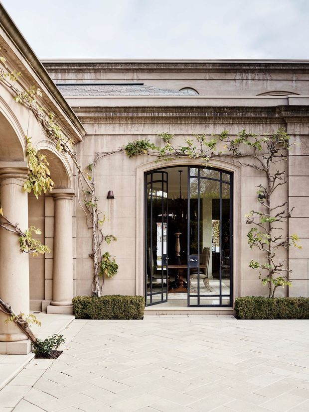 A refined stone-clad courtyard facade with classical architectural detailing, including arched openings, columns, layered cornices, and a tall black-framed glazed door. Climbing vines and clipped hedges soften the formal elevation, while pale rectangular paving creates a light, elegant courtyard setting. Through the glazing, a furnished interior is faintly visible.