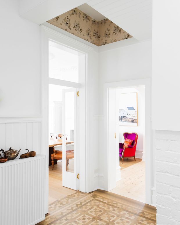 Bright white traditional hallway or entry passage with decorative timber parquet flooring, white timber wall panelling and moulded door surrounds. Two open doorways lead to adjacent furnished rooms, including a dining area and a room with a vivid pink-red armchair. A floral-patterned recessed ceiling panel adds a distinctive decorative feature, while a white-painted brick fireplace or wall edge is visible at right. A radiator and small tea-set display sit along the left wall.