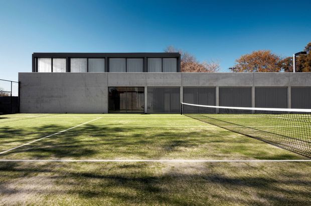 A modern low-rise building viewed from an outdoor tennis court. The structure has a long, rectilinear form with a smooth dark grey concrete or rendered facade, a black-framed clerestory-like upper level with vertical screened glazing, and a recessed central entrance. A tennis net and marked court dominate the foreground, with mature trees visible behind the building.