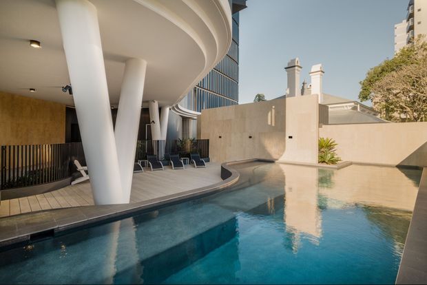 A sophisticated outdoor pool courtyard beside a contemporary building, featuring a curving pool, broad overhanging roof canopy supported by sculptural white columns, dark vertical screening, timber-toned wall panels, and a pale rendered boundary wall with classical chimney forms. Sun loungers line the pool edge, and the water reflects the surrounding architecture.