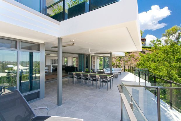 A bright, contemporary multi-level outdoor entertaining terrace with a large dining table, lounge seating, glass balustrades and expansive glazing beneath a projecting upper floor. Pale tiled paving, white rendered surfaces and dark-framed windows overlook leafy surroundings.