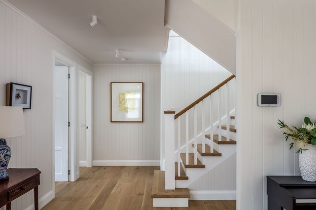 Bright coastal-style hallway and stairway with white vertical wall panelling, light timber flooring, and a white staircase with warm timber treads and handrail. A framed artwork, console furniture, indoor plants, and a wall-mounted control panel add detail to the airy interior.