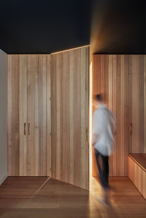 A dramatic, minimalist interior dominated by full-height vertical timber cabinetry and panelled doors. A partially open pivoting timber door reveals a warmly lit passage or adjoining space, while a blurred moving person adds a sense of motion. The dark ceiling contrasts with the warm wood-lined walls and timber flooring, creating a refined, sculptural composition.