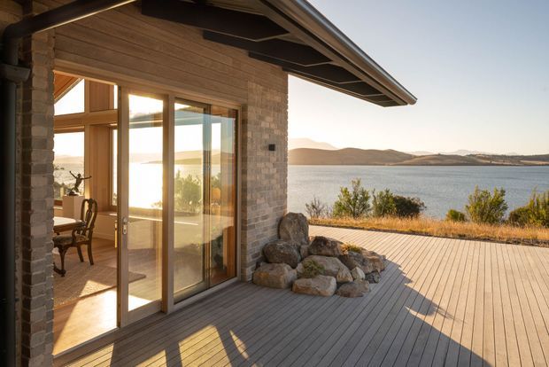 A lakeside residential exterior viewed from a broad timber deck. The building has a warm masonry wall, extensive timber-framed glazing and a projecting pitched roof with exposed eaves. Through the open glass doors, a dining area is visible. Large natural rocks border the deck, with calm water and low hills beyond.