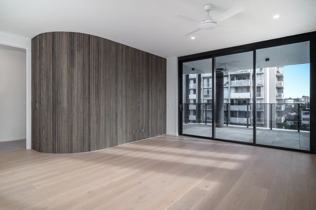 Bright, empty contemporary apartment living area with pale timber flooring, a curved full-height dark timber feature wall with concealed cabinetry, and expansive black-framed sliding glass doors opening to a balcony. A white ceiling fan and recessed lighting complete the clean, minimal interior, with neighbouring apartment buildings visible outside.