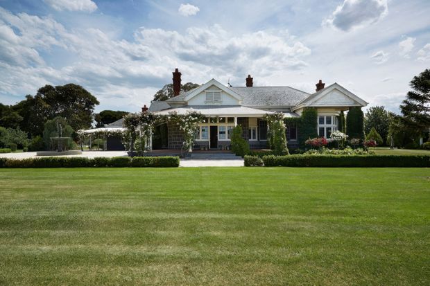A single-storey traditional house viewed from a broad manicured lawn. The light-coloured weatherboard facade has multiple gabled roof sections, white decorative trims, brick chimneys, a covered front verandah with climbing roses, and symmetrical garden planting. Mature trees and carefully maintained hedges frame the residential setting.
