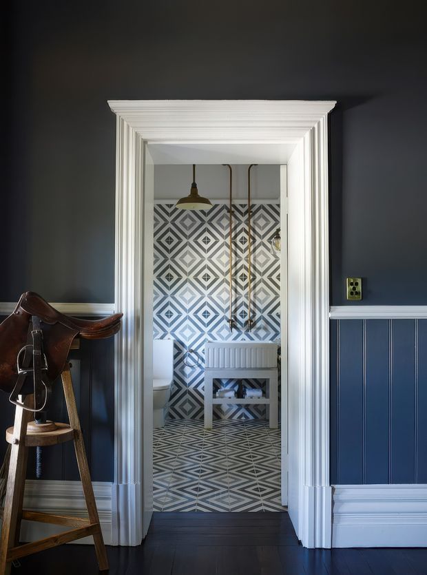 A dark, refined hallway or entry area with navy-blue wall panelling, white decorative mouldings and trim, and dark timber flooring. An open doorway leads to a compact bathroom featuring bold geometric patterned wall and floor tiles, a white radiator, exposed plumbing, and a pendant light. A vintage wooden saddle stand occupies the left foreground.