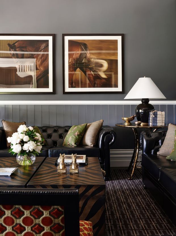 A richly styled living room with charcoal-grey walls, dark vertical wall panelling, framed artwork, and a pair of black tufted leather sofas and armchairs. A sculptural black table lamp, patterned cushions, an ornate coffee table, floral arrangement, and layered geometric rugs create a dramatic, sophisticated interior.