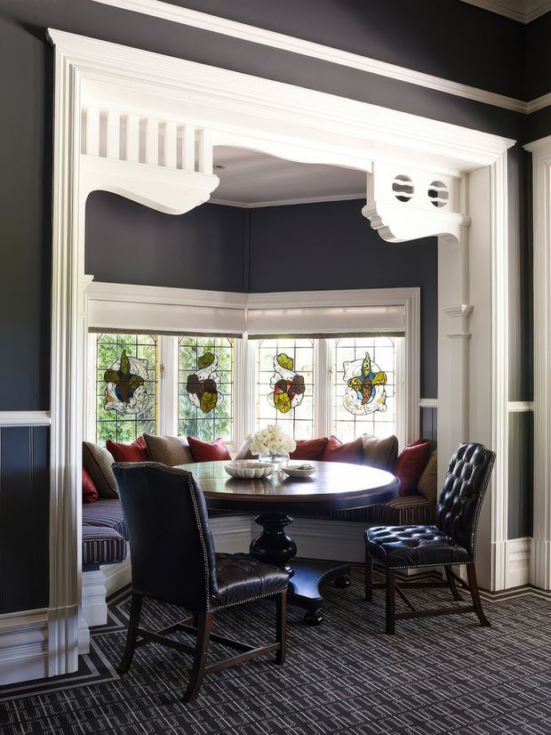 A refined dining nook with a round dark wood dining table, upholstered dining chairs, and built-in banquette seating beneath a curved bay window. The room features dramatic dark navy walls, extensive white painted architectural trim, decorative plaster or timber arch detailing, stained-glass window panels, and patterned carpet. The overall character is formal, historic, and richly detailed.