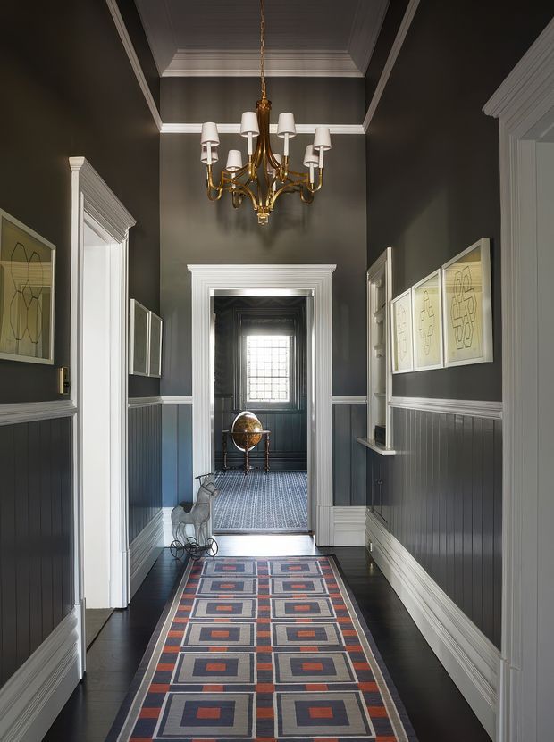 A narrow, elegant hallway with charcoal-grey walls, white decorative trim and wainscoting, dark timber flooring, and a long geometric runner rug. A traditional brass multi-arm chandelier hangs from a coffered ceiling. Framed artwork lines the walls, and the hallway opens to a secondary room with a patterned carpet and a bright leaded-glass window.