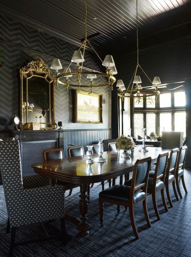 A dramatic, formal dining room with a large polished dark timber dining table surrounded by upholstered dining chairs and a patterned armchair. The room features dark chevron-patterned wallpaper, dark vertical wall panelling, a black slatted ceiling, ornate gilded mirrors, a dark mantel or sideboard, framed artwork, and two large brass-and-white pendant chandeliers. Tall multi-pane windows provide strong natural light, creating a refined, high-contrast traditional interior.