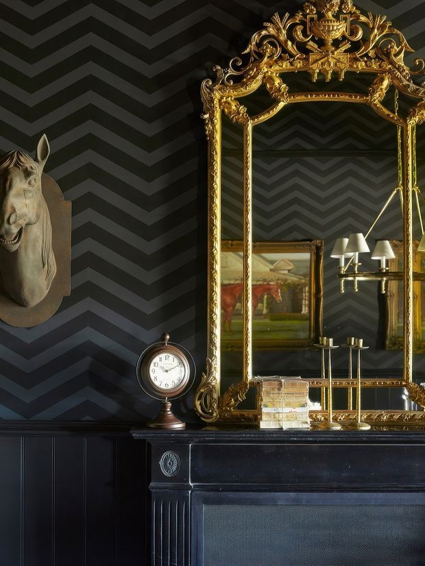 A moody, highly decorative interior vignette featuring a large ornate gilded mirror above a dark timber fireplace mantel. The walls are covered in a bold charcoal and black chevron-patterned finish, with a classical horse-head wall sculpture, antique mantel clock, framed artwork reflected in the mirror, candleholders, and a table lamp. The composition has a dramatic, richly layered period-inspired character.