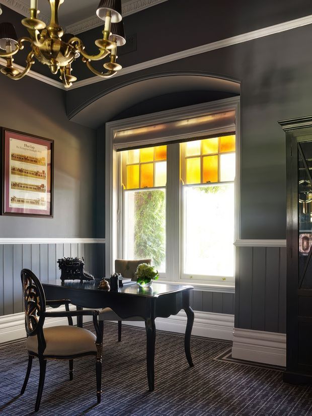 Elegant, moody study or writing room with dark charcoal walls, white decorative cornices and dado trim, blue-grey vertical wall panelling, patterned dark carpet, and a classic black writing desk with an upholstered chair. A brass chandelier, framed artwork, stained-glass transom panels, and a tall dark built-in cabinet create a refined heritage-inspired interior.