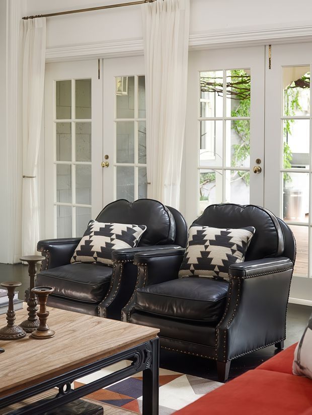 A refined sitting room with two black leather wingback armchairs, patterned monochrome cushions, and a substantial wood-and-metal coffee table. White French doors with divided glazing, full white curtains, decorative wall mouldings, and brass hardware bring abundant daylight and a classic architectural character. A geometric area rug and red upholstered seating add contrast.