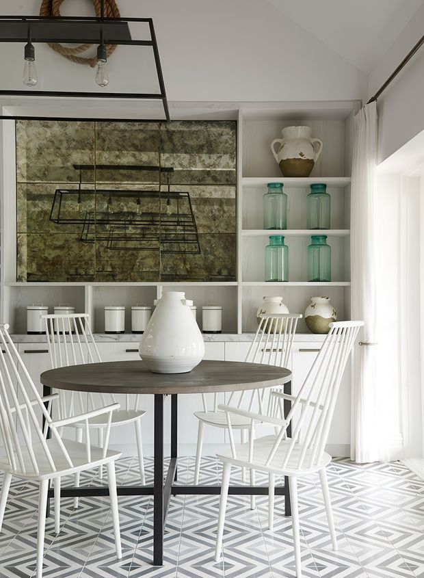 Bright, refined dining area with a round dark wood dining table and white spindle-back chairs. The room features built-in white shelving and cabinetry displaying ceramic vessels and translucent green glass jars, a large mottled stone or marble-look feature backsplash, geometric patterned floor tiles, sheer curtains, and black geometric pendant lighting. The palette is predominantly white, grey, black, and muted green, creating a crisp contemporary yet eclectic interior.