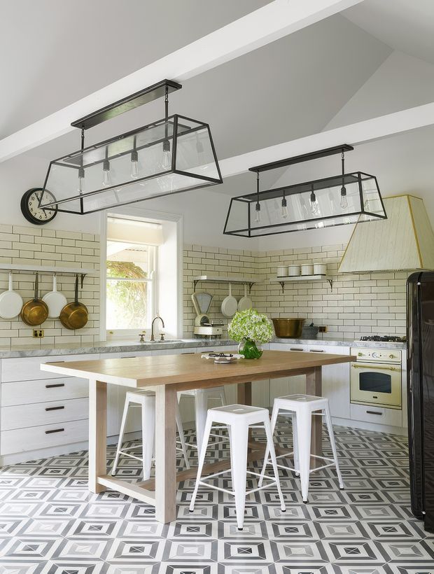 Bright, spacious kitchen with white cabinetry, white subway-tile walls, a patterned geometric tile floor, and a large timber island with white metal stools. Two oversized black-framed pendant lights hang from a coffered or beamed white ceiling. Open shelving displays crockery, while copper pans hang above the stone benchtop. A freestanding cream-coloured range cooker and a large sculptural rangehood feature on the right.