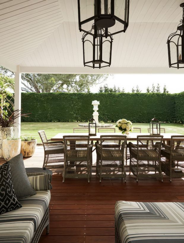 A covered outdoor dining and lounge area with a white-lined pitched ceiling, dark timber decking, woven dining chairs, striped upholstered seating, and large black lantern pendant lights. The open terrace overlooks a manicured lawn, clipped hedge, and formal garden feature, creating a refined garden entertaining space.